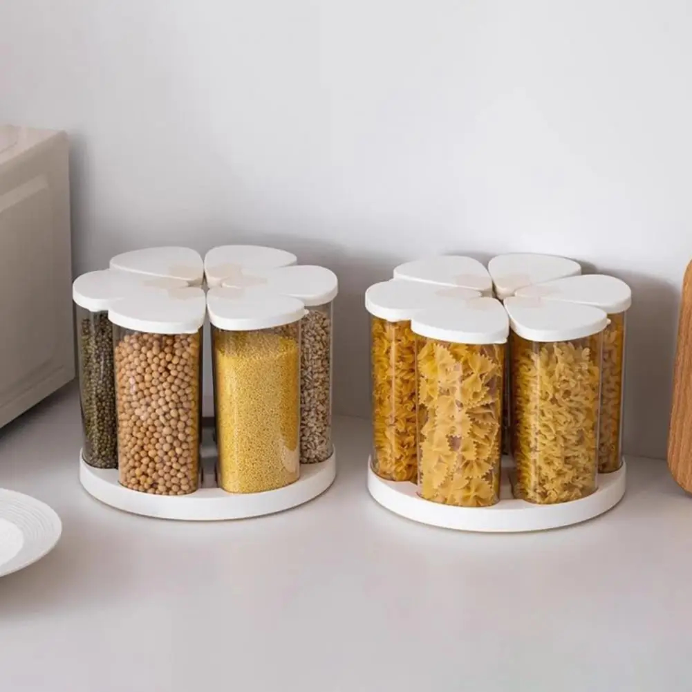 Two white flower-shaped spice jars with clear cylindrical containers and white lids, filled with dried beans and pasta, displayed on a light countertop against a white wall.