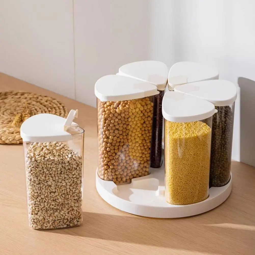 A white round spice jar organizer with four clear cylindrical containers filled with different grains and seeds, shown on a light wood tabletop. A matching single clear container with a white lid stands beside the organizer.