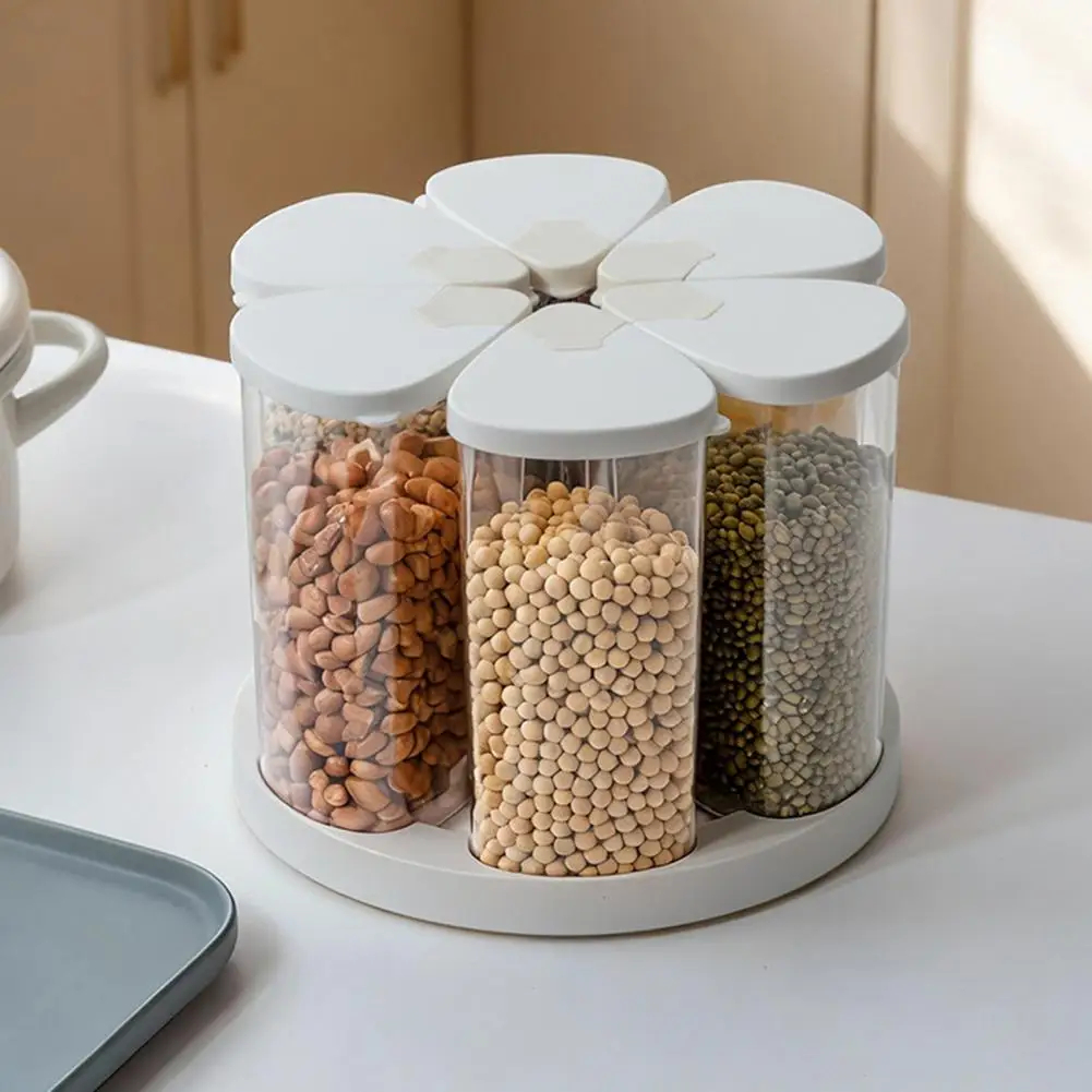 A flower-shaped rotating spice and dry goods container set with three clear cylindrical jars filled with peanuts, beans, and lentils, on a white tabletop.
