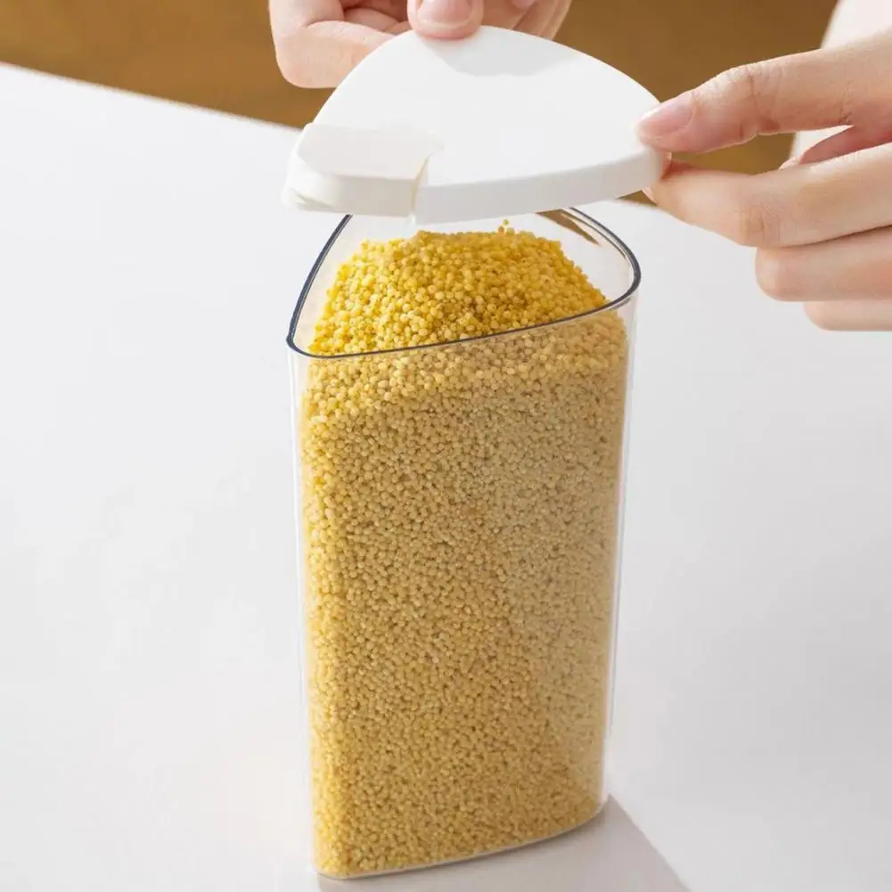 A clear plastic storage container filled with yellow grain or millet, shown on a white surface with a person's hands lifting a white triangular lid.