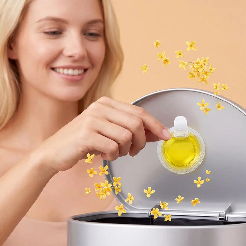 A woman inserts a small round yellow oil capsule into a silver appliance, with small yellow flower graphics floating around the product against a warm beige background.